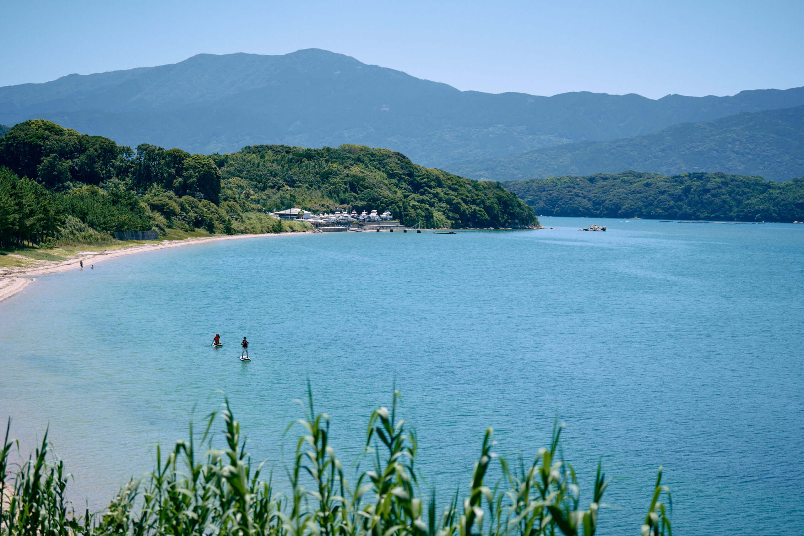 透明感のある青い海でSUPを楽しむ人々と、背景に広がる緑豊かな山々の風景