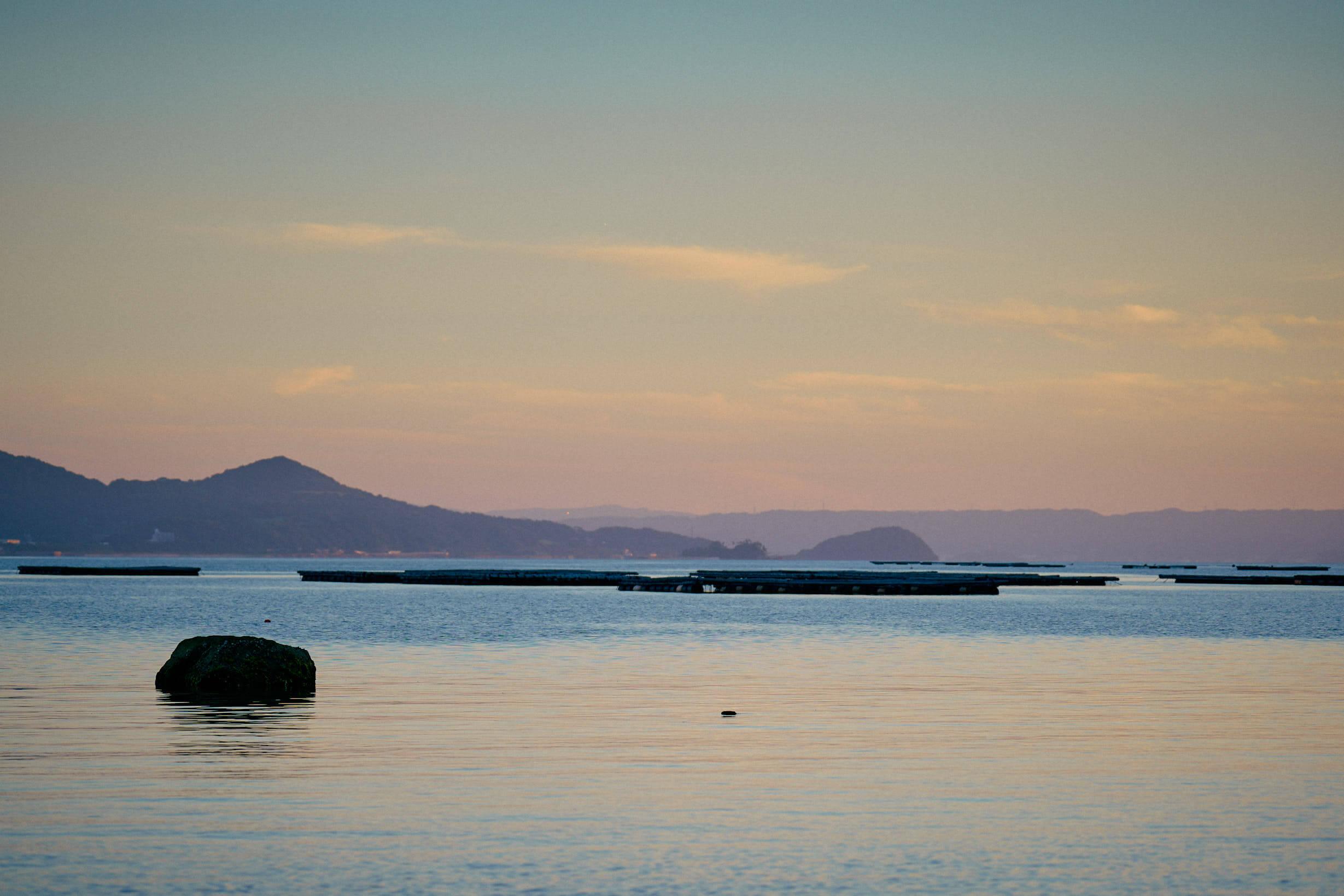 夕暮れ時の穏やかな海。手前に岩が浮かび、遠くには漁場のいかだと山並みがシルエットで映る